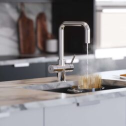 A D-shape water tap in chrome runs hot water over uncooked pasta in a pot