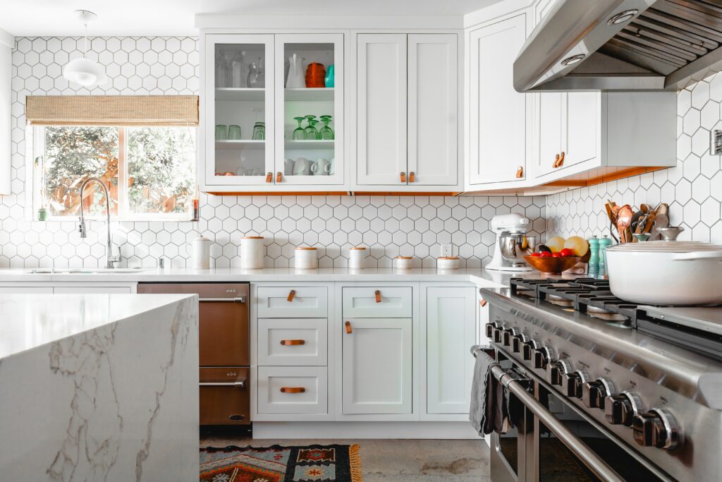 an elegant white kitchen cabinets with flame orange handles, stainless steel range cooker and a sliding window in front of sink with a chrome swan neck tap