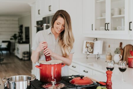 a smiling woman standing and putting pepper on a stock pot 